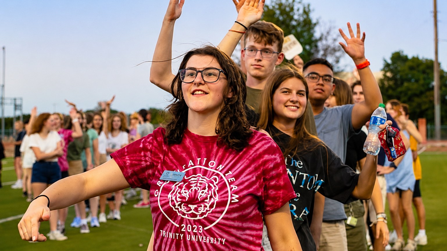Orientation leaders and first-year students raising their hands and cheering in a line on the athletic field at dusk